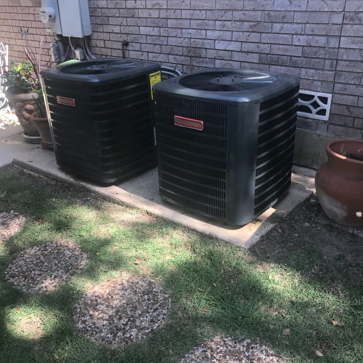 Two Goodman condensing units side by side on a pad along a brick wall