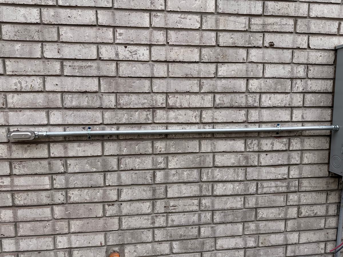 New galvanized steel conduit strapped across a brick wall from a service enclosure to a pulling elbow, with new conductors not yet landed at the enclosure