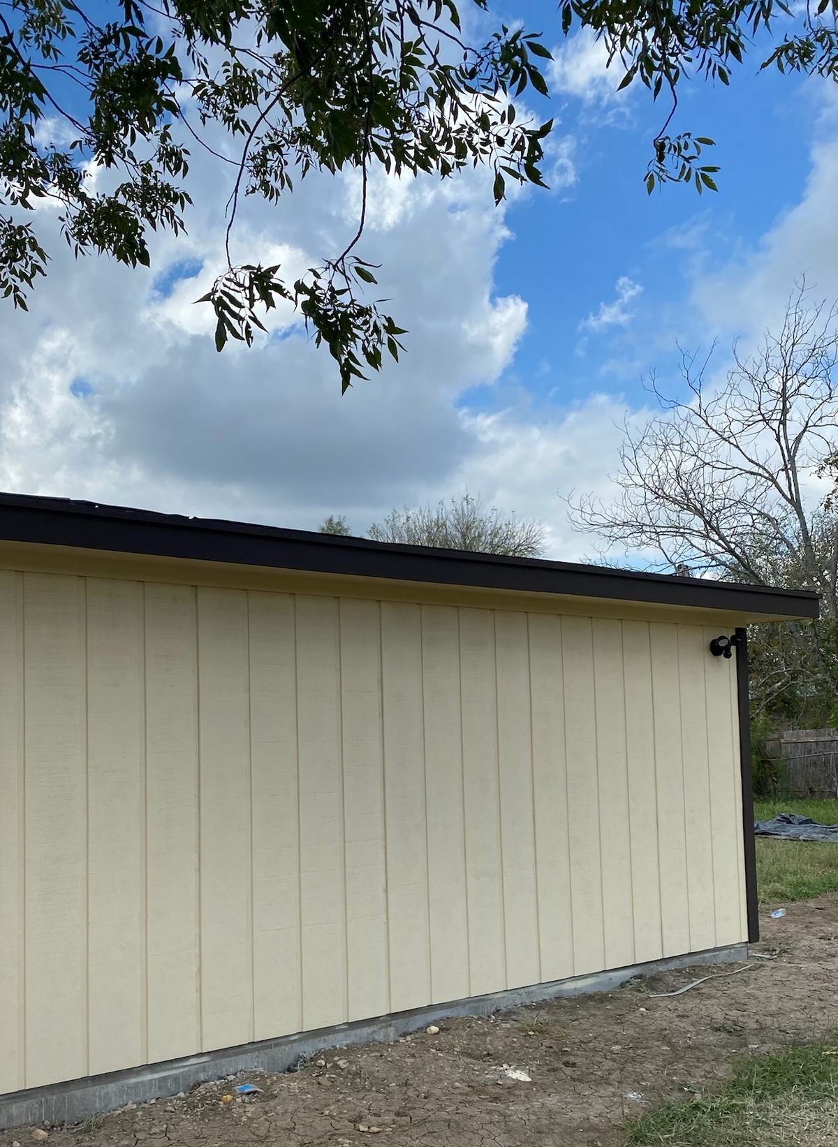 Long side wall of a room addition with cream vertical siding and dark trim above bare soil