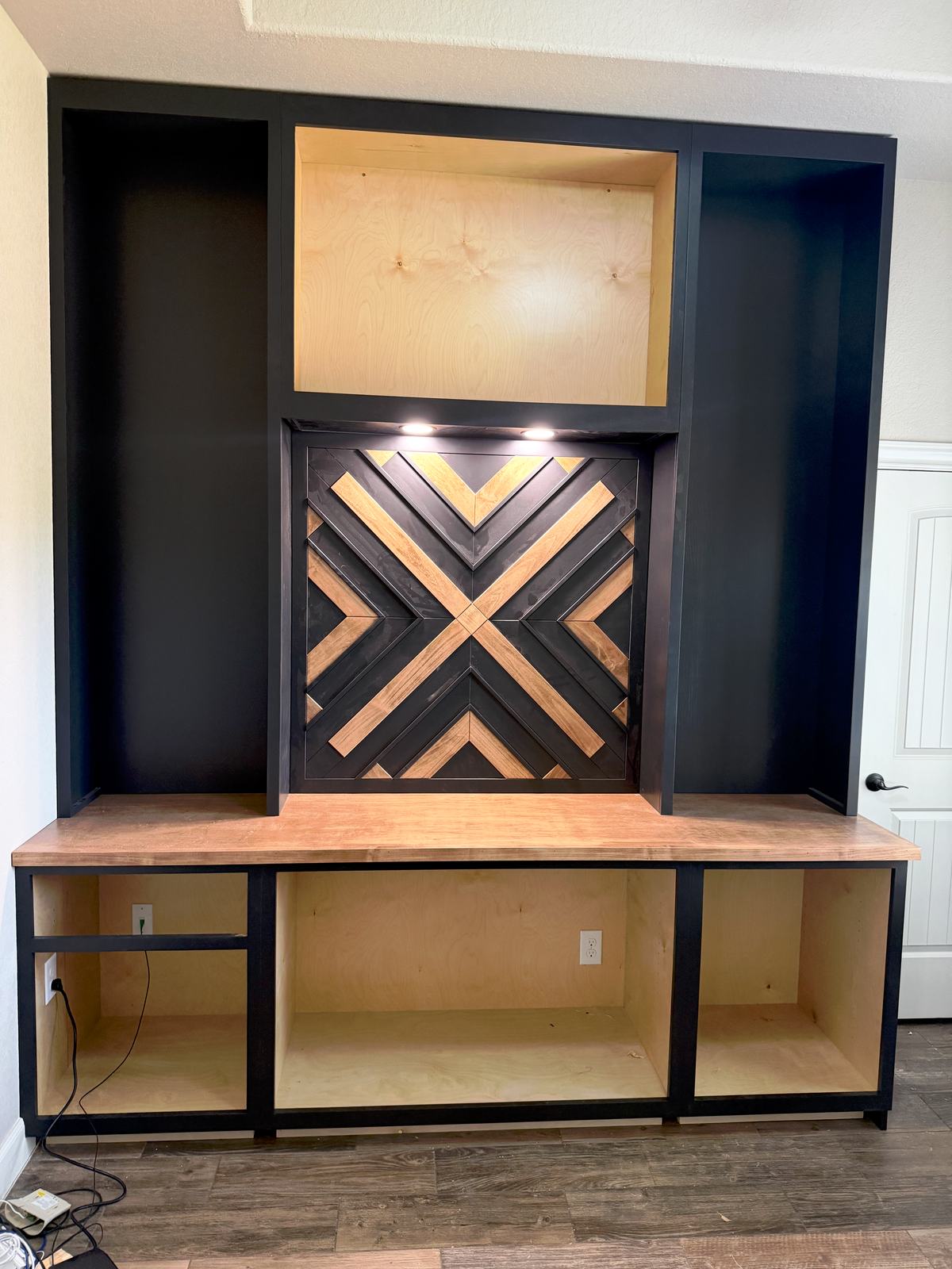Charcoal built-in wall unit under construction with a chevron wood center panel under puck lights and open plywood bays