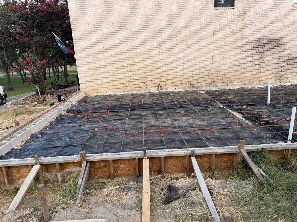 Rebar grid over a vapor barrier inside wood slab forms beside a brick wall, ready for the pour