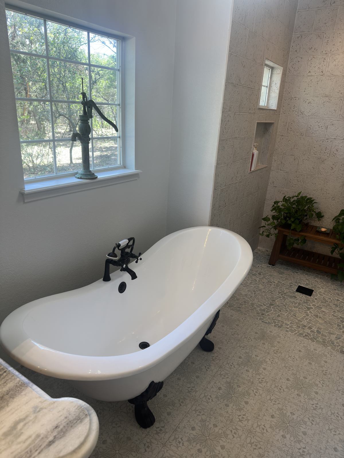 White clawfoot tub with a dark wall-mounted faucet on a patterned tile floor under a window