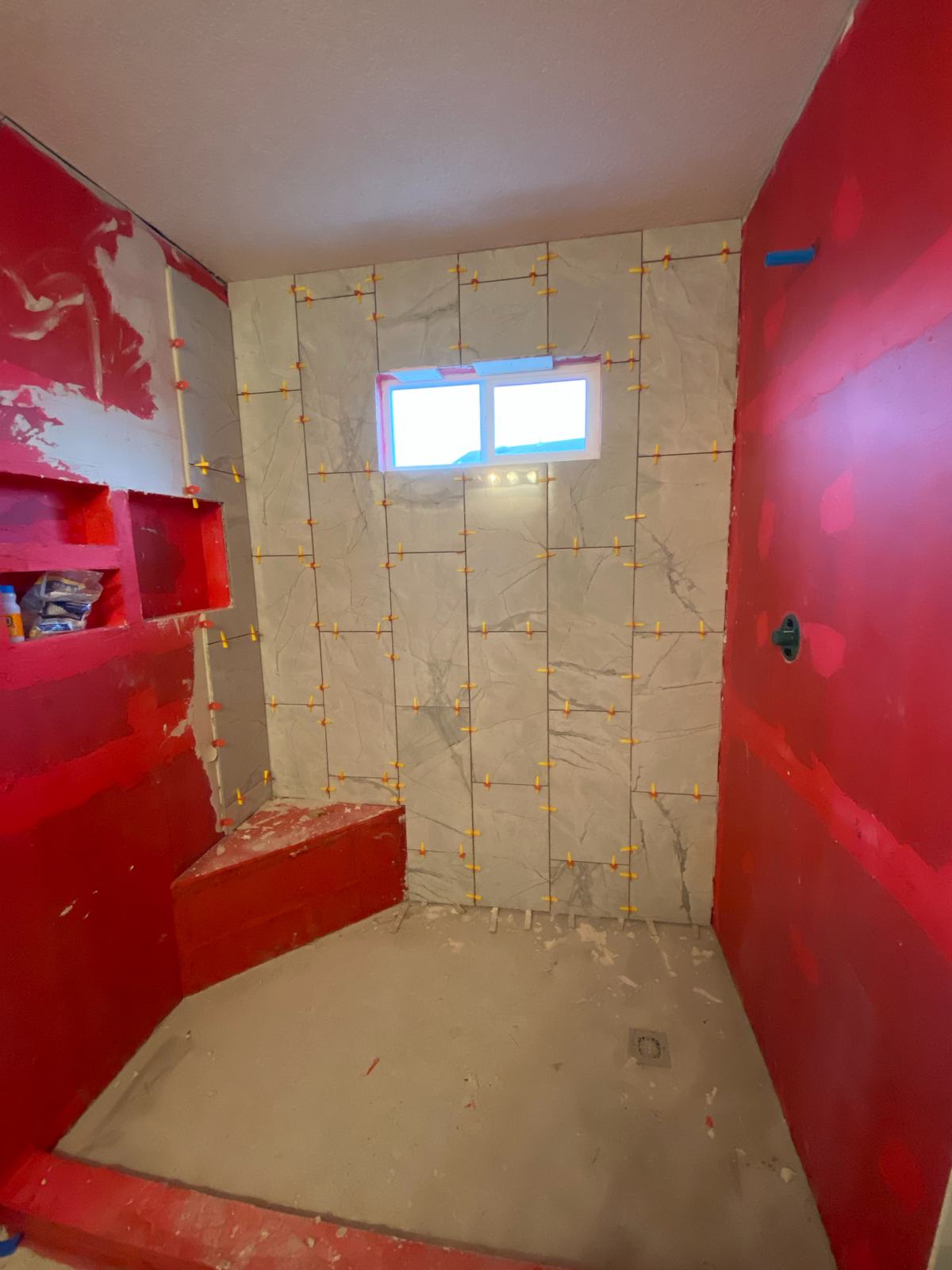 Shower walls being tiled over red waterproofing membrane, with leveling clips on the new tile and a bench formed