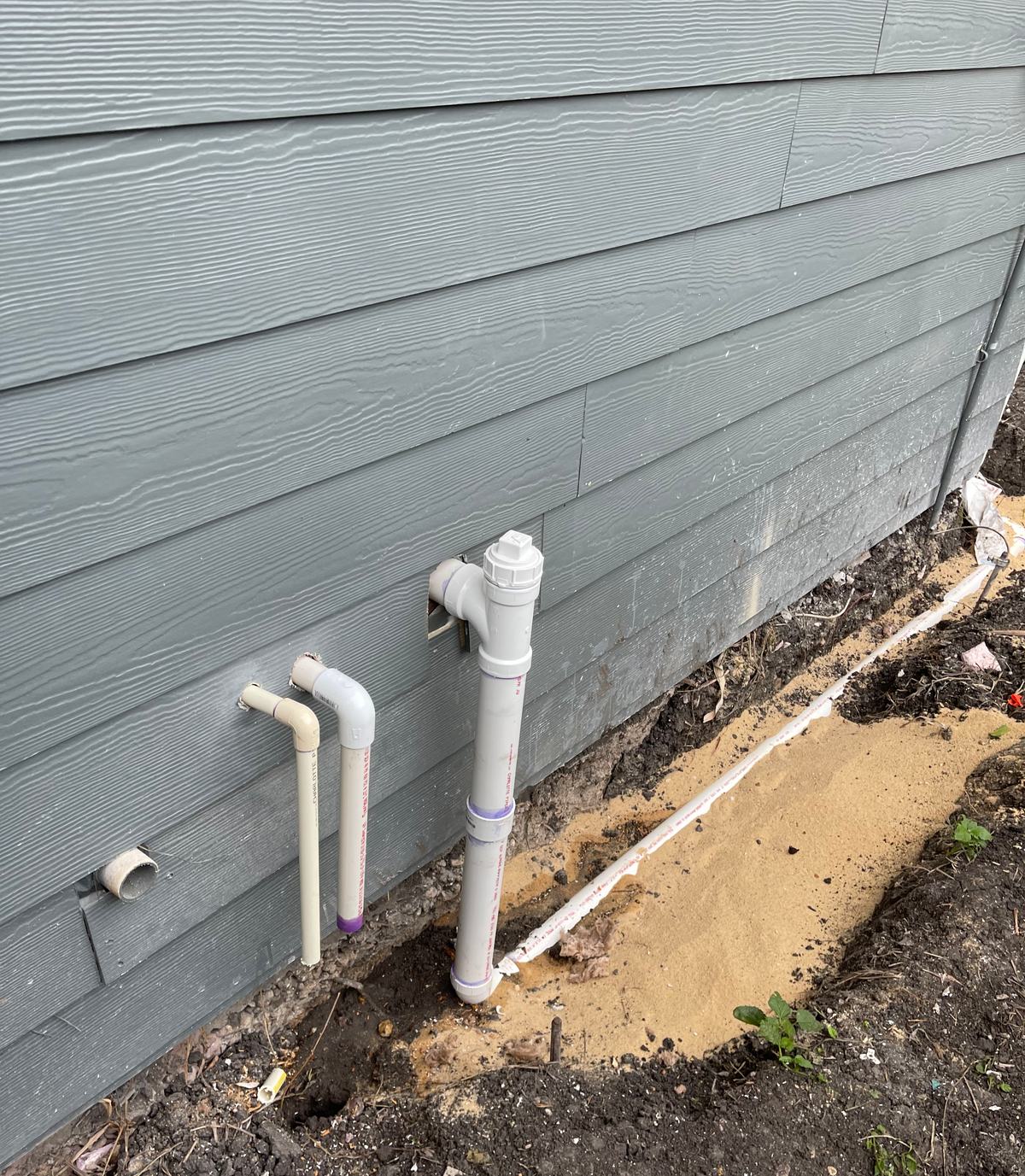 New PVC cleanout riser beside two smaller PVC lines along a house wall above sand-bedded trench