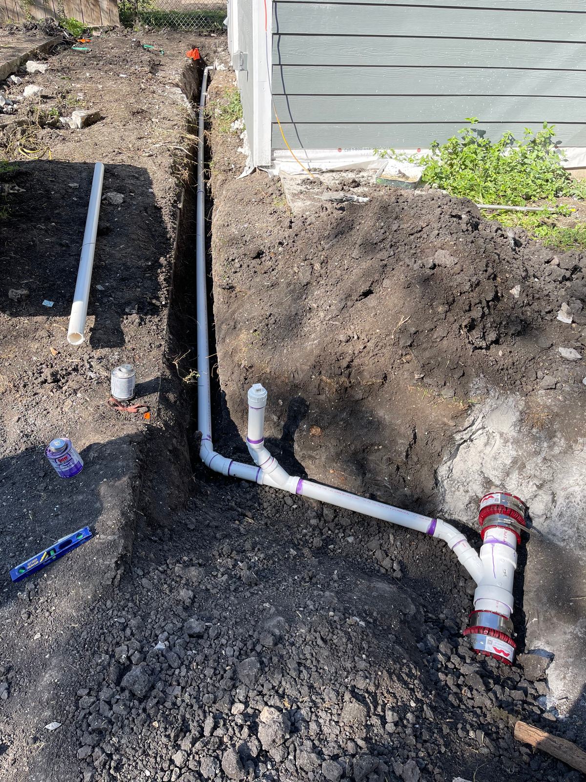 New PVC sewer line laid in an open trench along a house with fittings and couplings at the tie-in